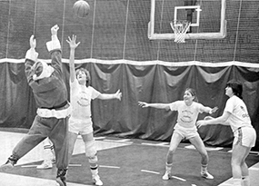 Santa Claus takes a jump shot while being guarded by one female basketball player while two other players defend and watch.